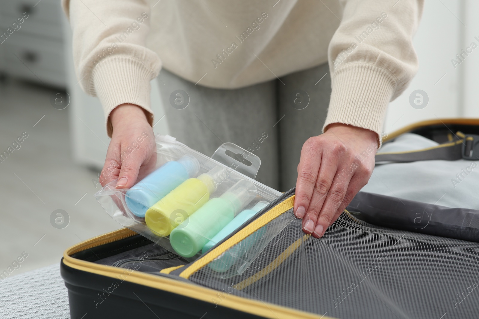 Photo of Woman with plastic bag of cosmetic travel kit packing suitcase at home, closeup. Bath accessories
