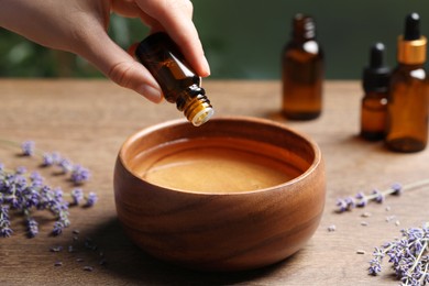 Photo of Woman dripping essential oil from bottle into bowl near lavender at wooden table, closeup