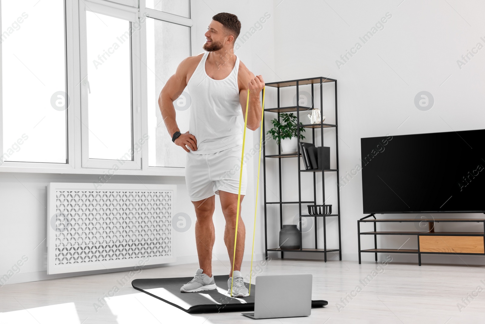 Photo of Muscular man doing exercise with elastic resistance band near laptop on mat at home