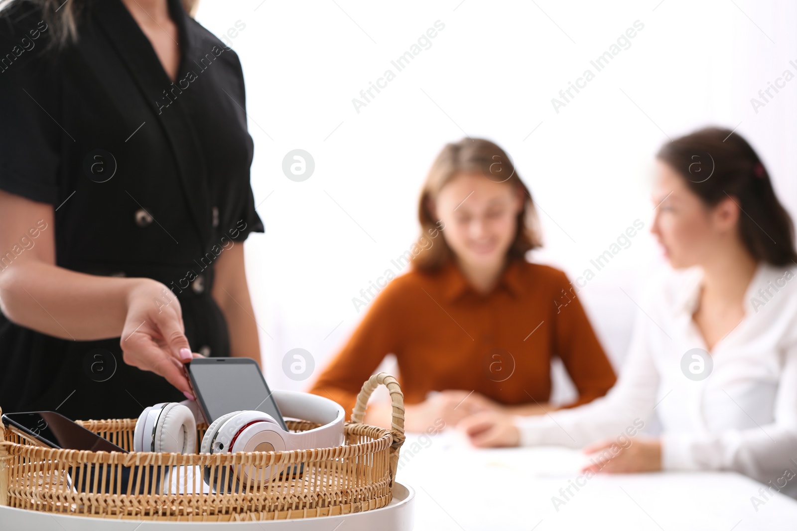 Photo of Woman putting smartphone into wicker basket with gadgets in office, closeup. Digital detox concept