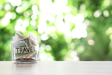 Photo of Glass jar with money and label TRAVEL on table against blurred background. Space for text