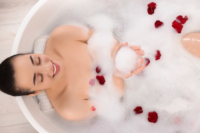 Happy woman taking bath in tub with foam and rose petals indoors, top view