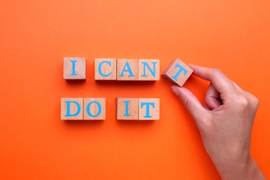 Photo of Motivation concept. Woman changing phrase from I Can't Do It into I Can Do It by removing wooden cube with letter T on orange background, top view
