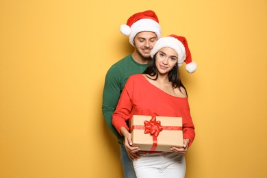 Photo of Young happy couple with Santa hats holding gift box on yellow background. Christmas celebration