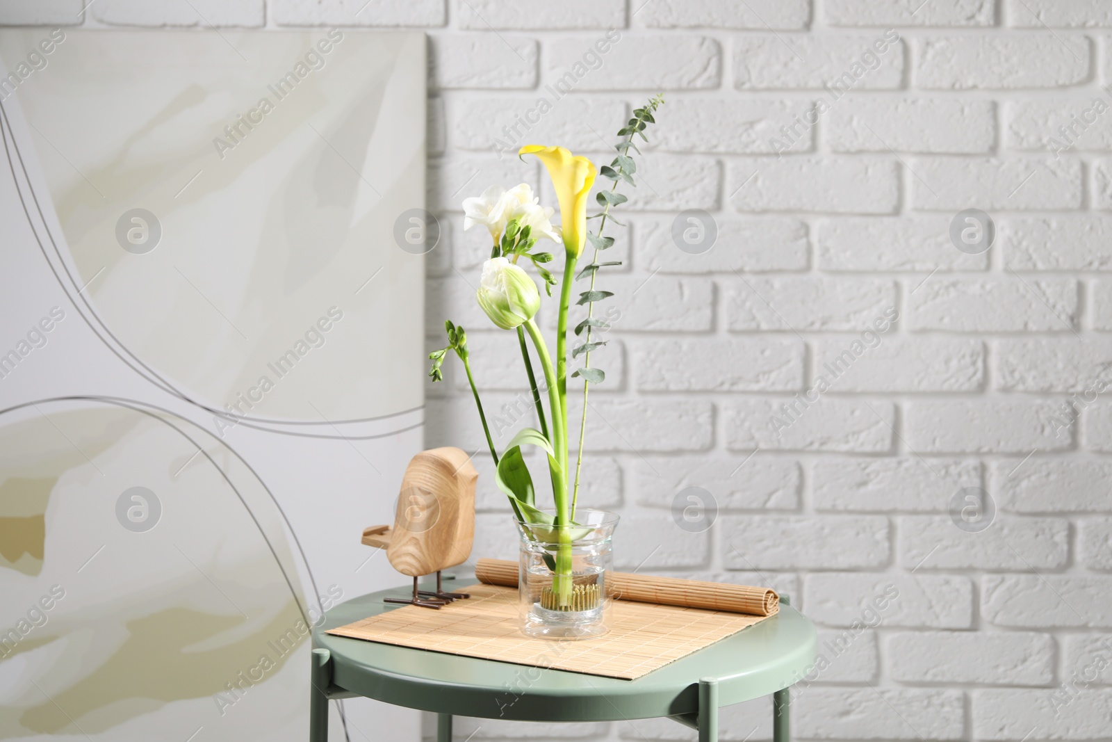 Photo of Beautiful ikebana for stylish house decor. Floral composition with fresh flowers and eucalyptus branch on coffee table near white brick wall