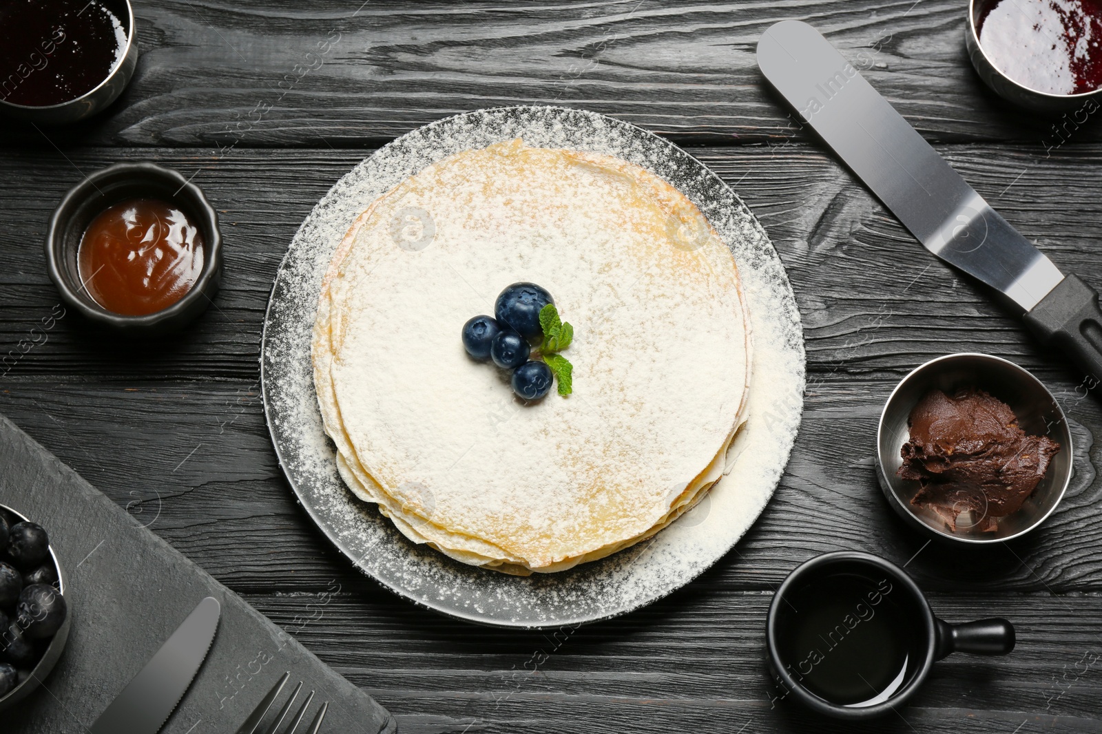 Photo of Delicious crepes with blueberries and different jams served on black wooden table, flat lay