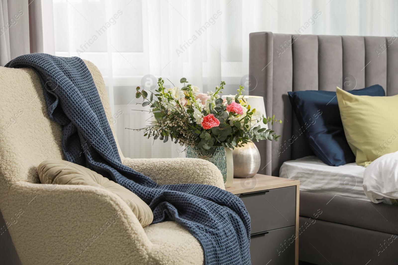 Photo of Armchair, lamp and beautiful bouquet on wooden bedside table near comfortable bed in room. Stylish interior