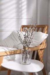 Photo of Glass vase with pussy willow tree branches on table near armchair in room