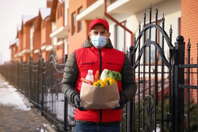 Photo of Courier in medical mask holding paper bag with groceries near house outdoors. Delivery service during quarantine due to Covid-19 outbreak