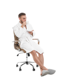 Handsome man with cup of coffee wearing bathrobe sitting in chair on white background