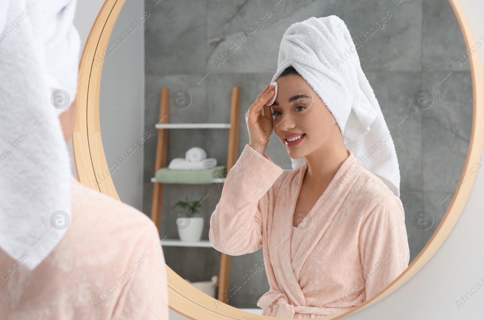 Photo of Beautiful woman in terry towel removing makeup with cotton pad near mirror indoors