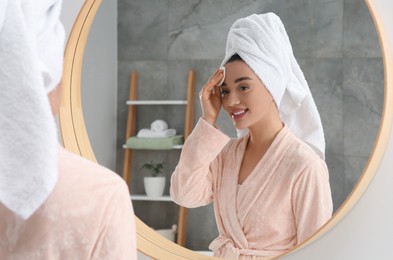 Photo of Beautiful woman in terry towel removing makeup with cotton pad near mirror indoors