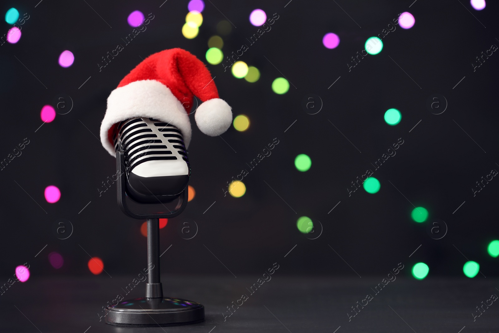 Photo of Retro microphone with Santa hat on table against blurred lights, space for text. Christmas music