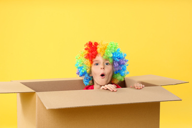 Photo of Little boy in clown wig sitting inside of cardboard box on yellow background. April fool's day