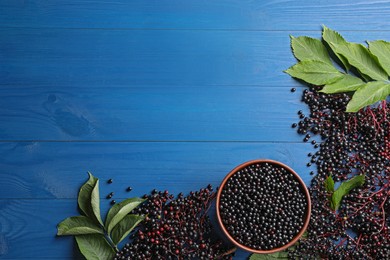 Photo of Ripe elderberries with green leaves on blue wooden table, flat lay. Space for text