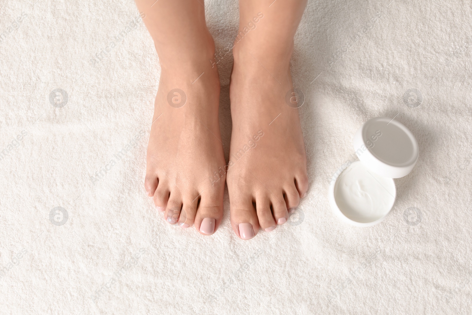 Photo of Woman with beautiful feet and jar of moisturizing cream on white towel, top view. Spa treatment