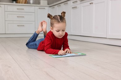 Photo of Cute little girl reading book on warm floor in kitchen, space for text. Heating system