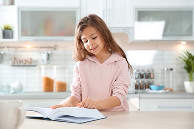 Photo of Cute little girl reading book at table in kitchen