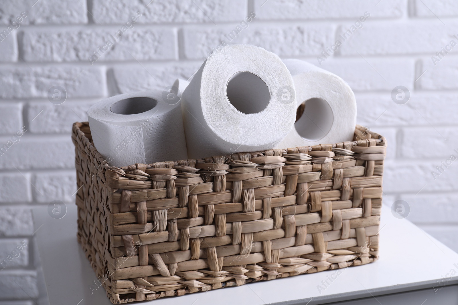 Photo of Toilet paper rolls in wicker basket on chest of drawers near white brick wall