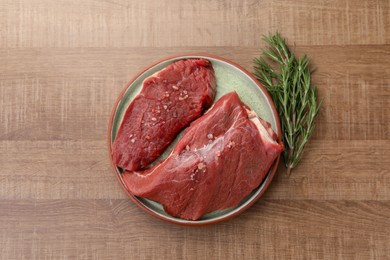 Photo of Pieces of raw beef meat with spices and rosemary on wooden table, flat lay