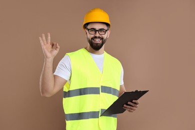 Photo of Engineer in hard hat holding clipboard and showing ok gesture on brown background