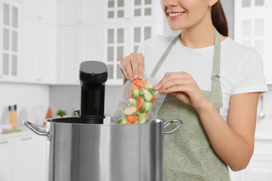 Woman putting vacuum packed vegetables into pot with sous vide cooker in kitchen, closeup. Thermal immersion circulator