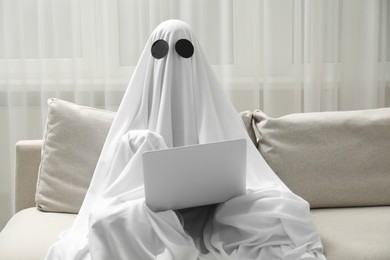 Photo of Creepy ghost. Person covered with white sheet using laptop on sofa at home