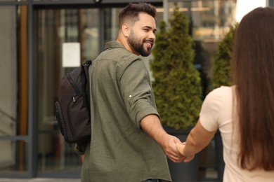 Photo of Long-distance relationship. Man with backpack holding hands with his girlfriend near building outdoors