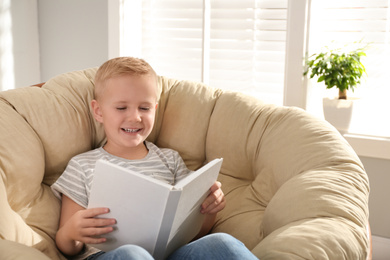 Photo of Cute little boy reading book in papasan chair at home
