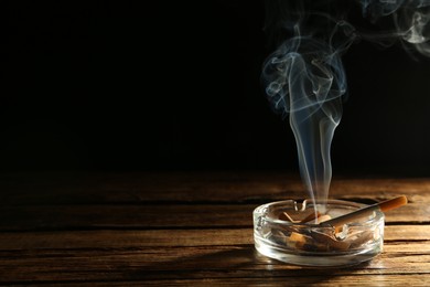 Smoldering cigarette in glass ashtray on wooden table against black background. Space for text