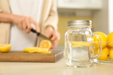 Photo of Woman cutting fruits in kitchen, focus on mason jar with lemon water