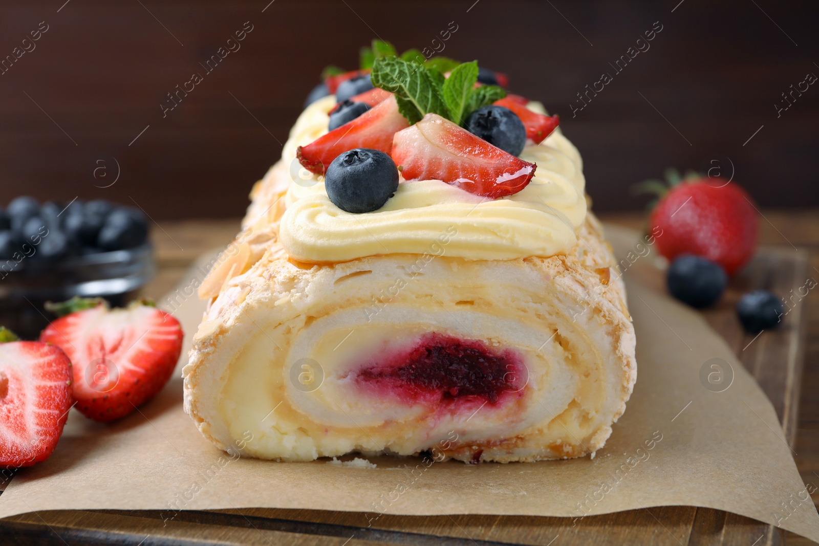 Photo of Tasty meringue roll with jam, cream, strawberry, blueberry and mint on wooden board, closeup