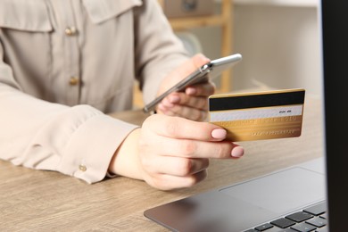 Online payment. Woman using smartphone and credit card near laptop at wooden table indoors, closeup