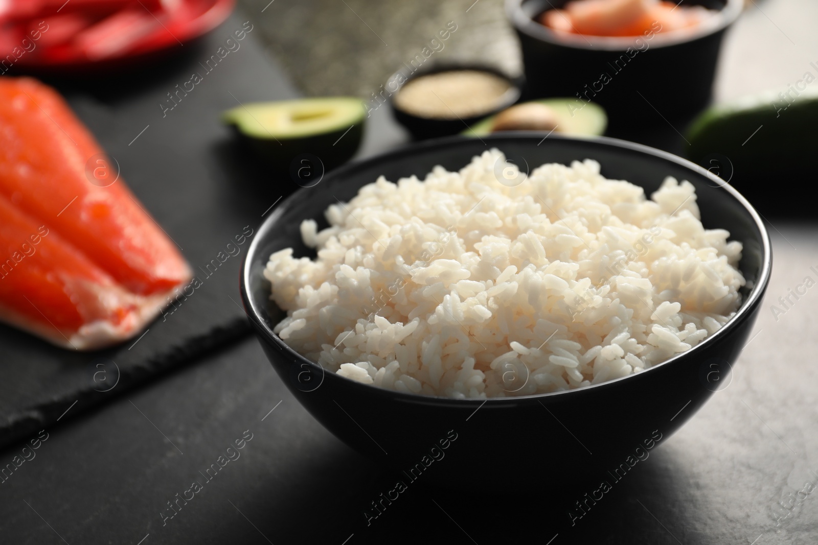 Photo of Cooked rice in bowl and other ingredients for sushi on dark table, closeup