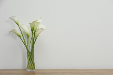 Photo of Beautiful calla lily flowers in glass vase on wooden table near white wall. Space for text
