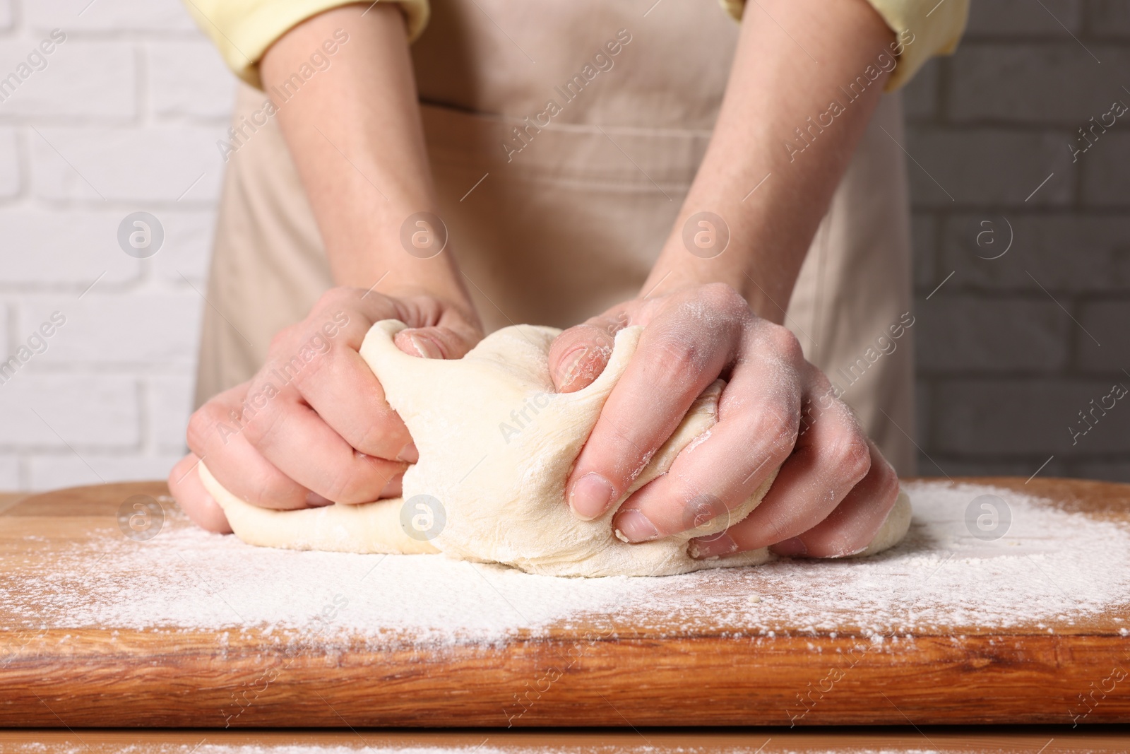 Photo of Woman kneading dough at wooden table near white brick wall, closeup