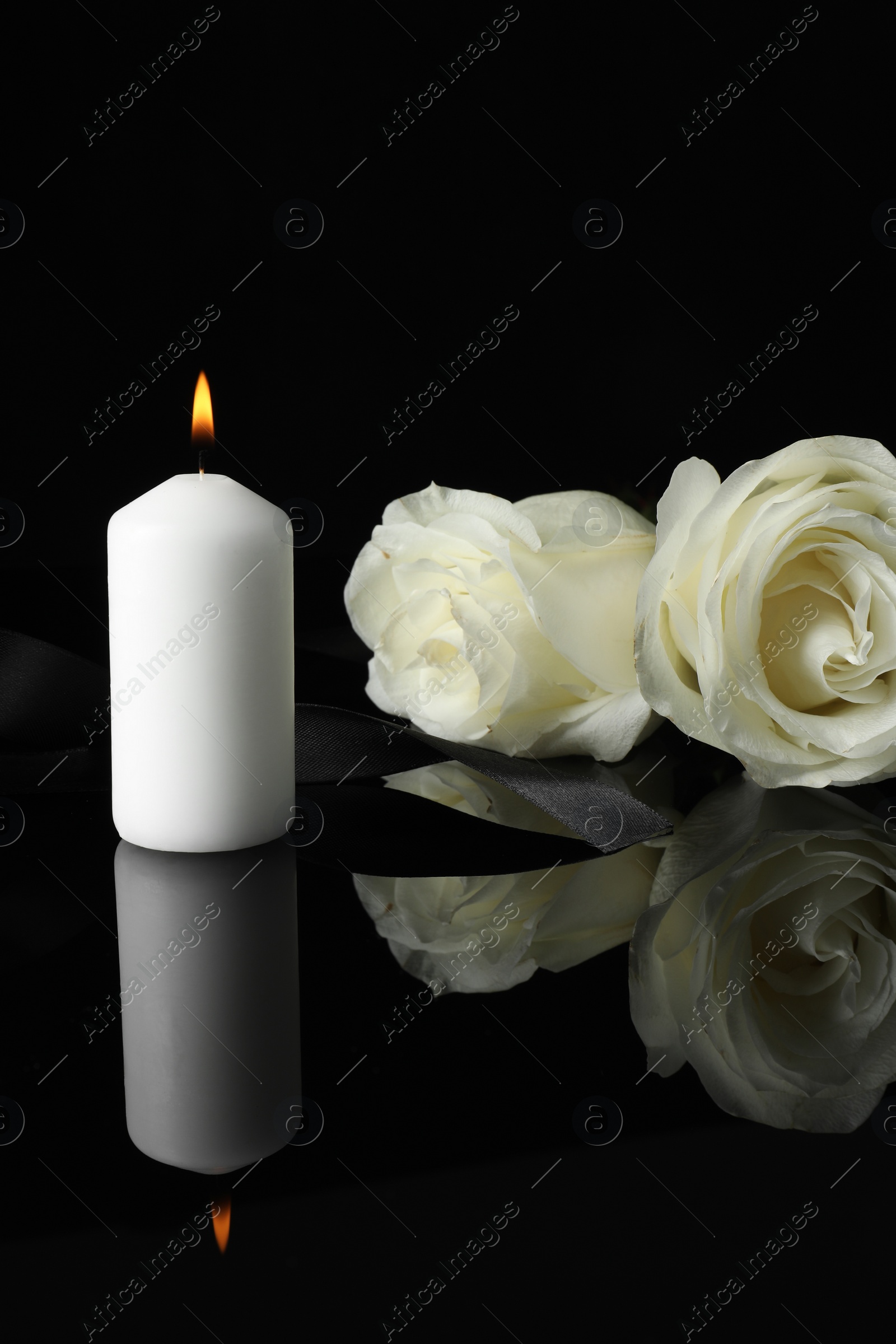 Photo of Burning candle, white roses and ribbon on black mirror surface in darkness, space for text. Funeral symbols