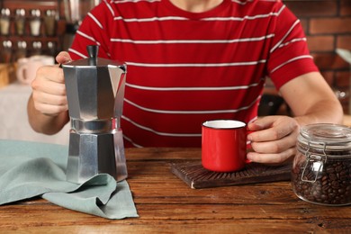 Brewing coffee. Man with jar of beans, moka pot and mug at wooden table indoors, closeup