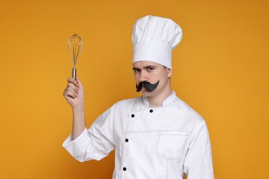 Photo of Portrait of happy confectioner with funny artificial moustache holding whisk on orange background