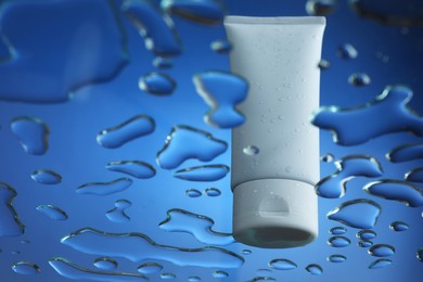Photo of Moisturizing cream in tube on glass with water drops against blue background, low angle view. Space for text