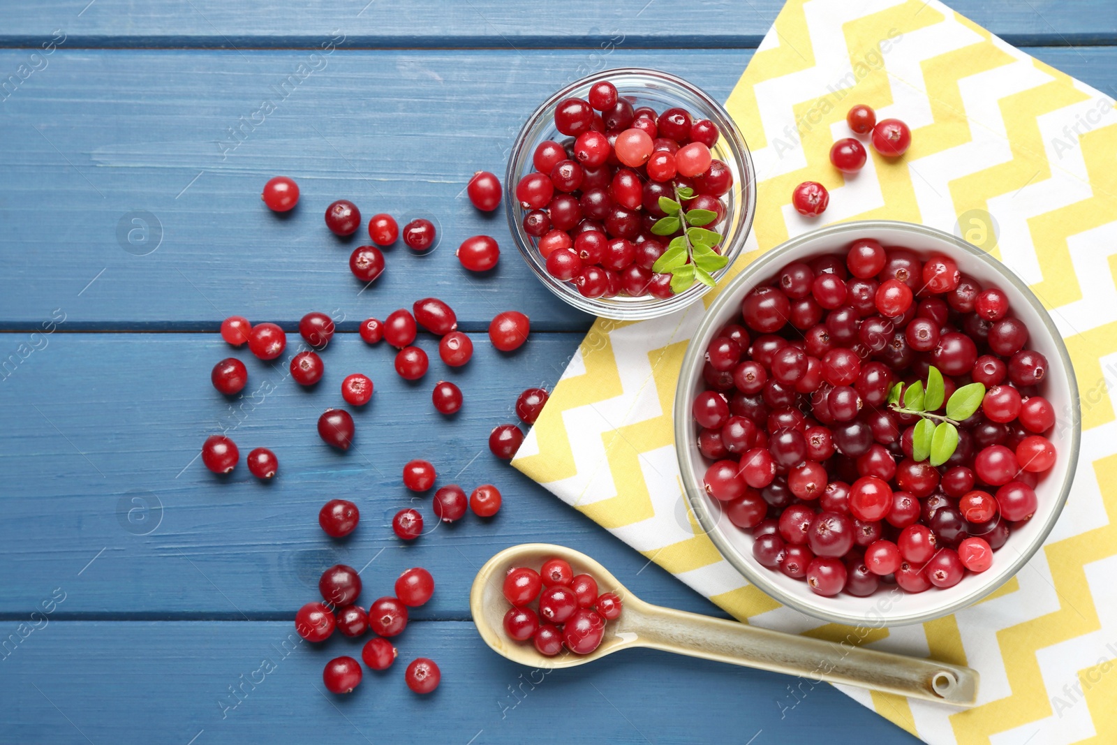 Photo of Fresh ripe cranberries in bowls and spoon on blue wooden table, flat lay