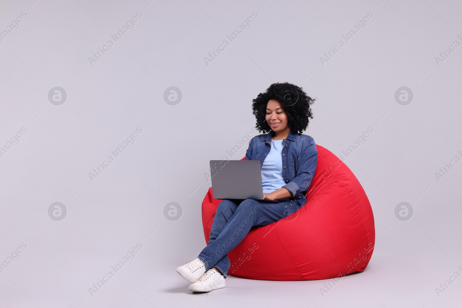 Photo of Happy young woman with laptop sitting on beanbag chair against light grey background. Space for text