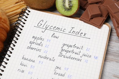 Notebook with products of low and moderate glycemic index, kiwi and chocolate on light wooden table, closeup
