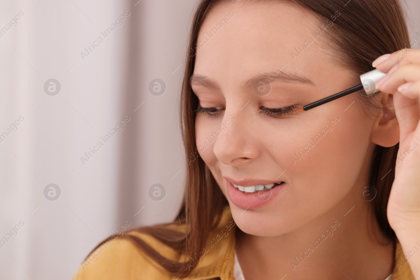 Photo of Beautiful woman applying serum onto eyelashes indoors, closeup