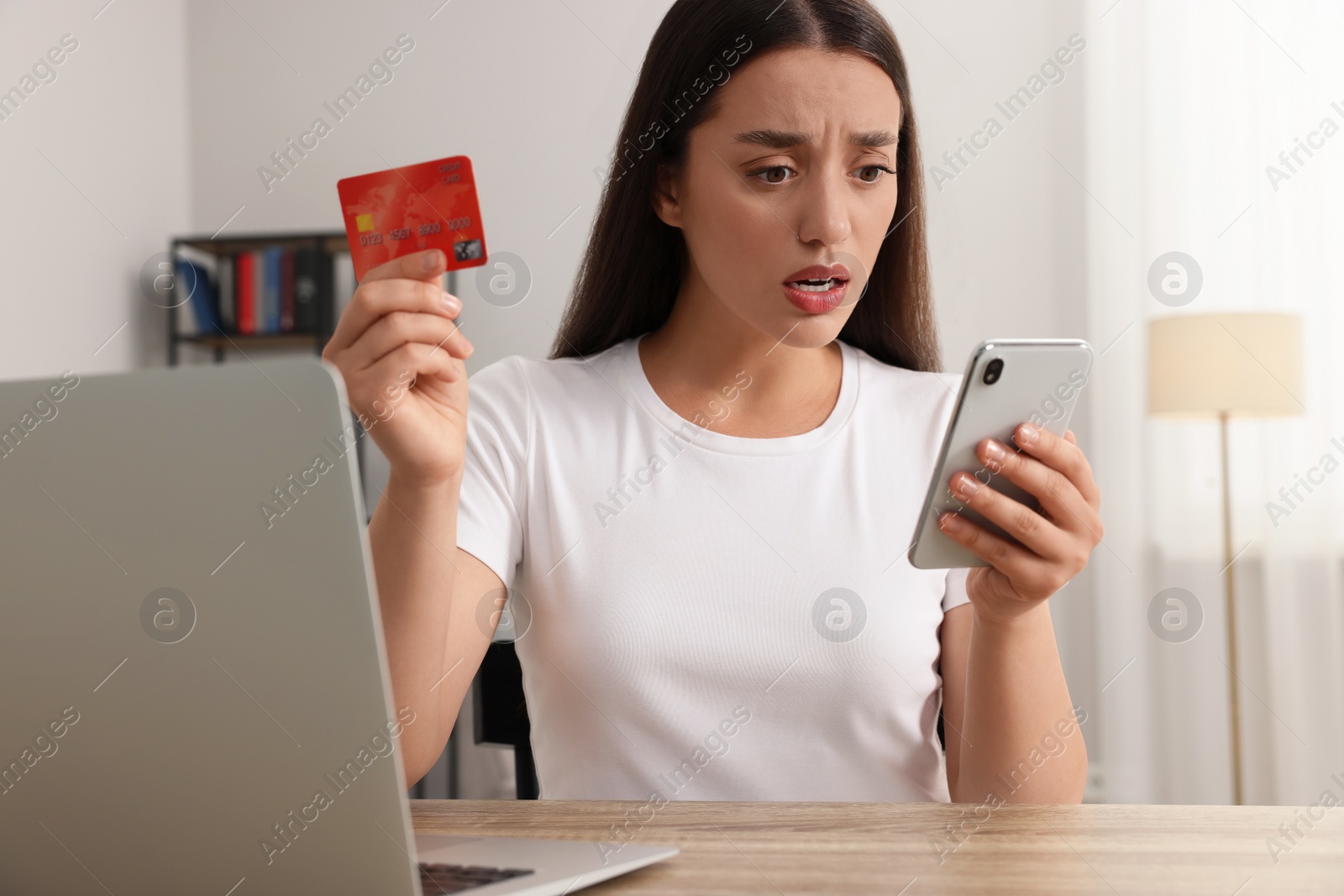 Photo of Shocked woman with credit card using smartphone and laptop at table indoors. Be careful - fraud