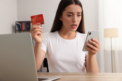 Shocked woman with credit card using smartphone and laptop at table indoors. Be careful - fraud