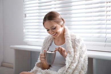 Beautiful young woman with cup of hot drink and marshmallow near window at home. Winter atmosphere