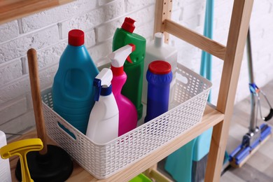 Photo of Plunger and basket with detergents on wooden shelf near white brick wall indoors