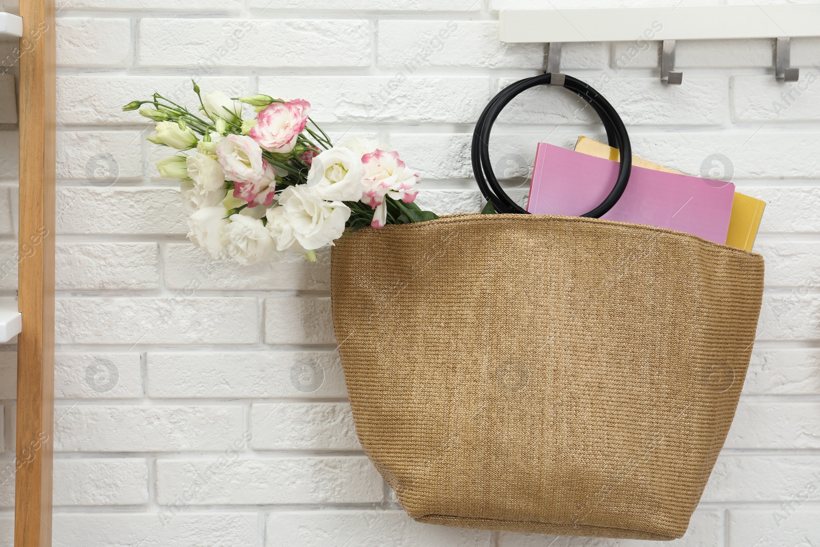 Photo of Stylish beach bag with beautiful bouquet and magazines hanging on hook rack indoors