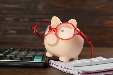 Calculator, notebook, pen and piggy bank with glasses on wooden table